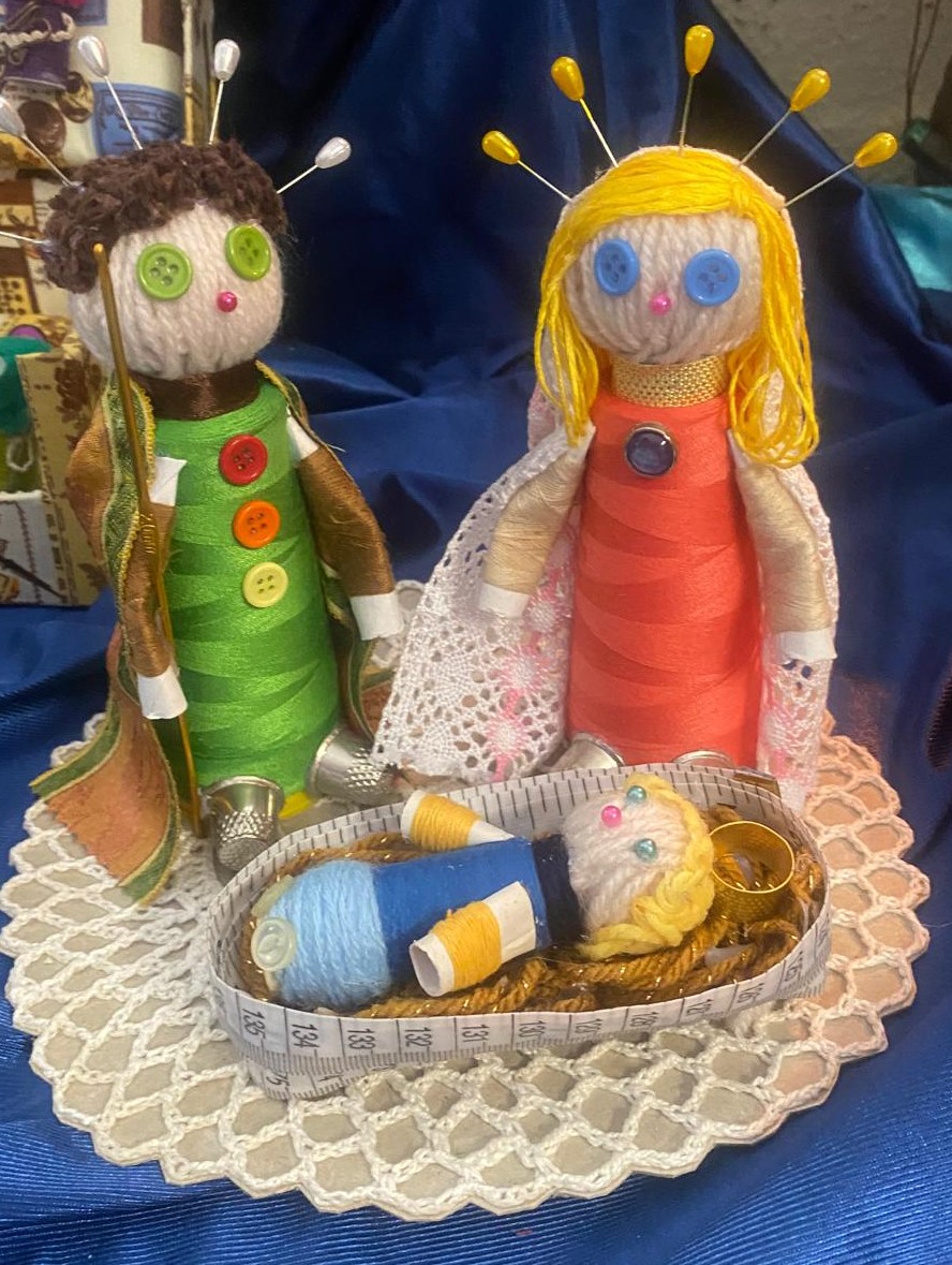Figuras de belén artesanales hechas con hilos y botones, representando a María, José y el niño Jesús en una cuna de papel de medir, sobre un fondo azul.