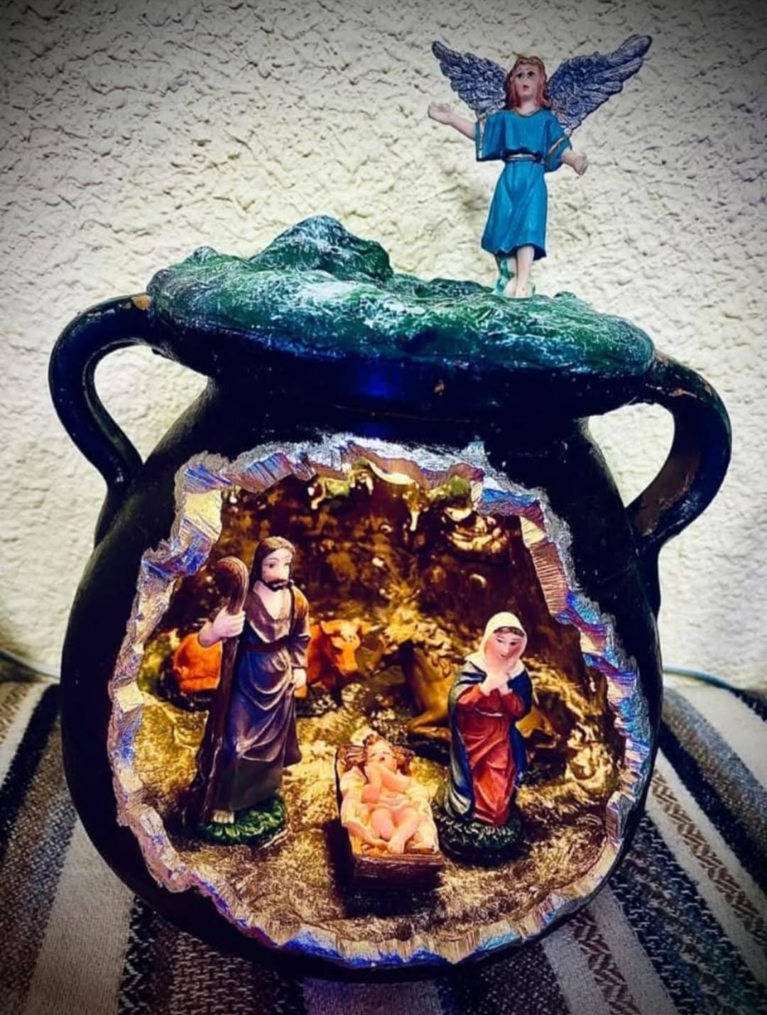 Representación de un belén artesanal dentro de un recipiente, con figuras del nacimiento de Jesús, incluyendo a María, José y el niño. En la parte superior, se destaca una figura de un ángel. El belén se presenta en un fondo decorativo.
